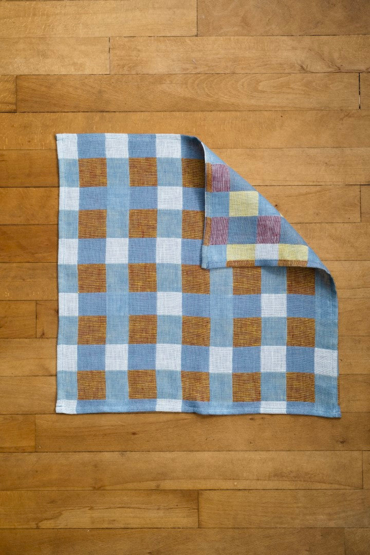 A checked linen tea towel laying on a wooden floor with one of its corners turned over to show the underside. 