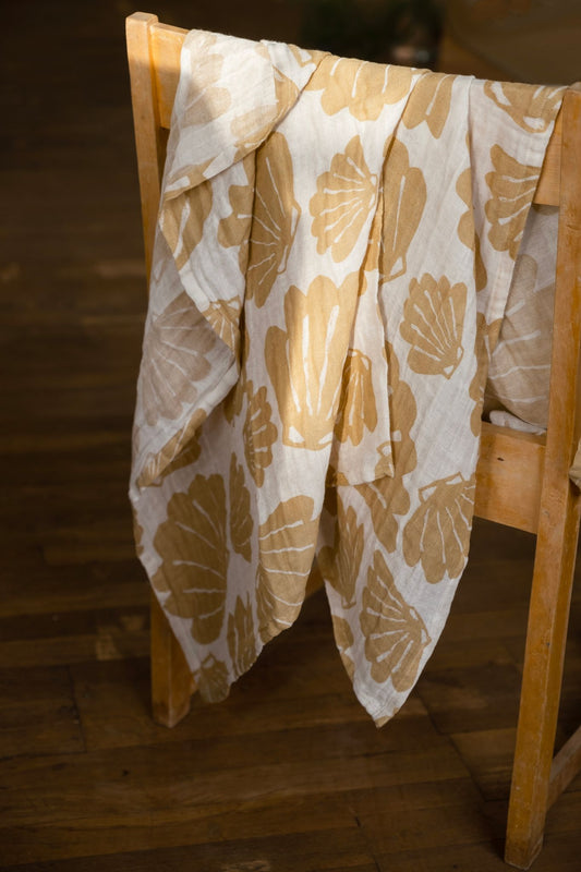 A muslin blanket with a shell pattern hanging on a wooden chair, predominantly in an off-white/sand colour.