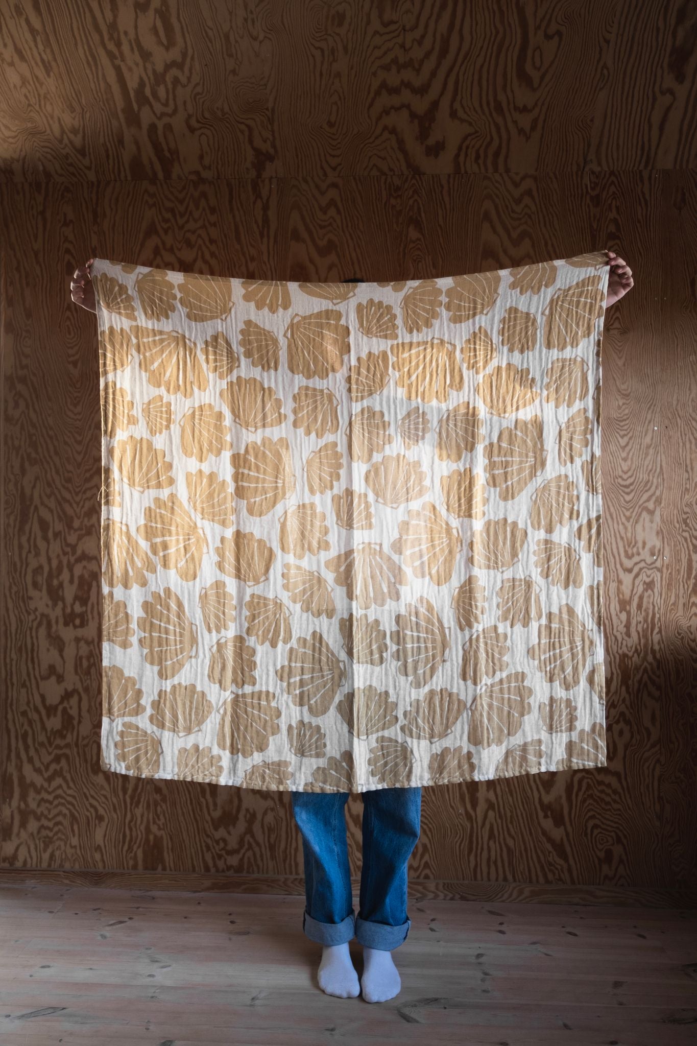 A person holding up an outstretched muslin blanket with a shell pattern in an off-white/sand colour.