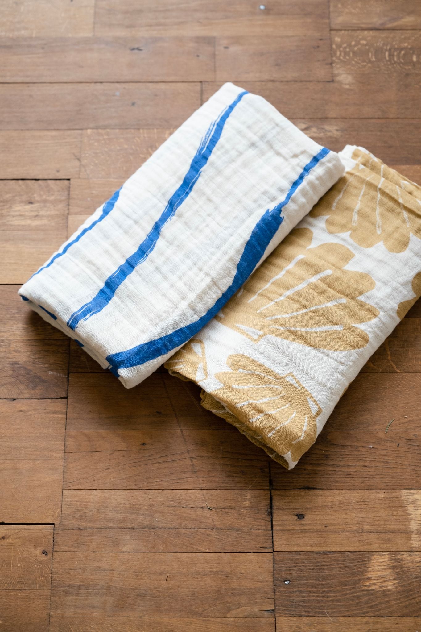 Two folded up muslin blankets, one in a blue and white colourway, the other in a sand and white colourway, on a wooden floor.