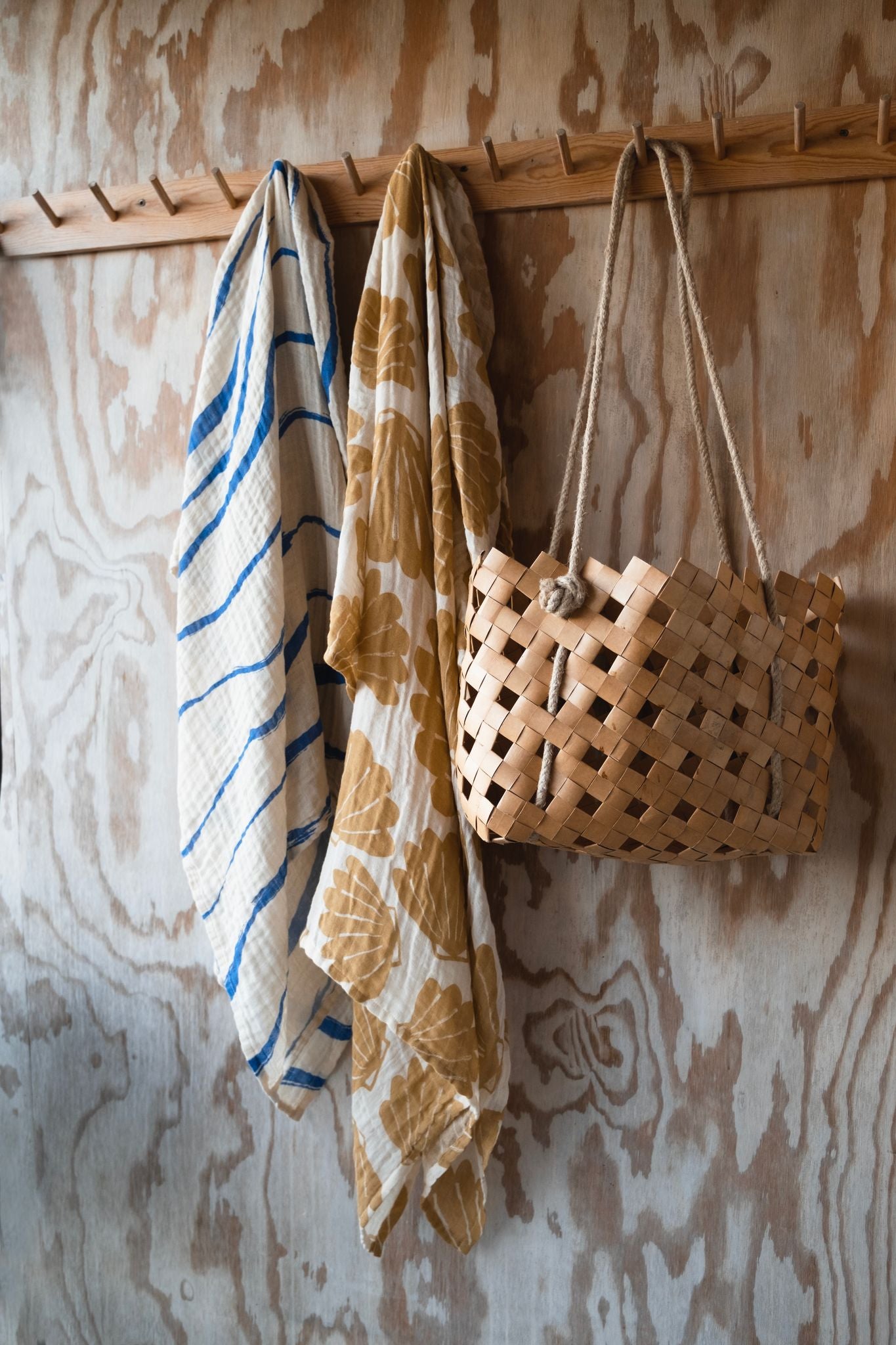 Two muslin clothes hanging off a wooden peg rail with a woven bag hanging next to them.