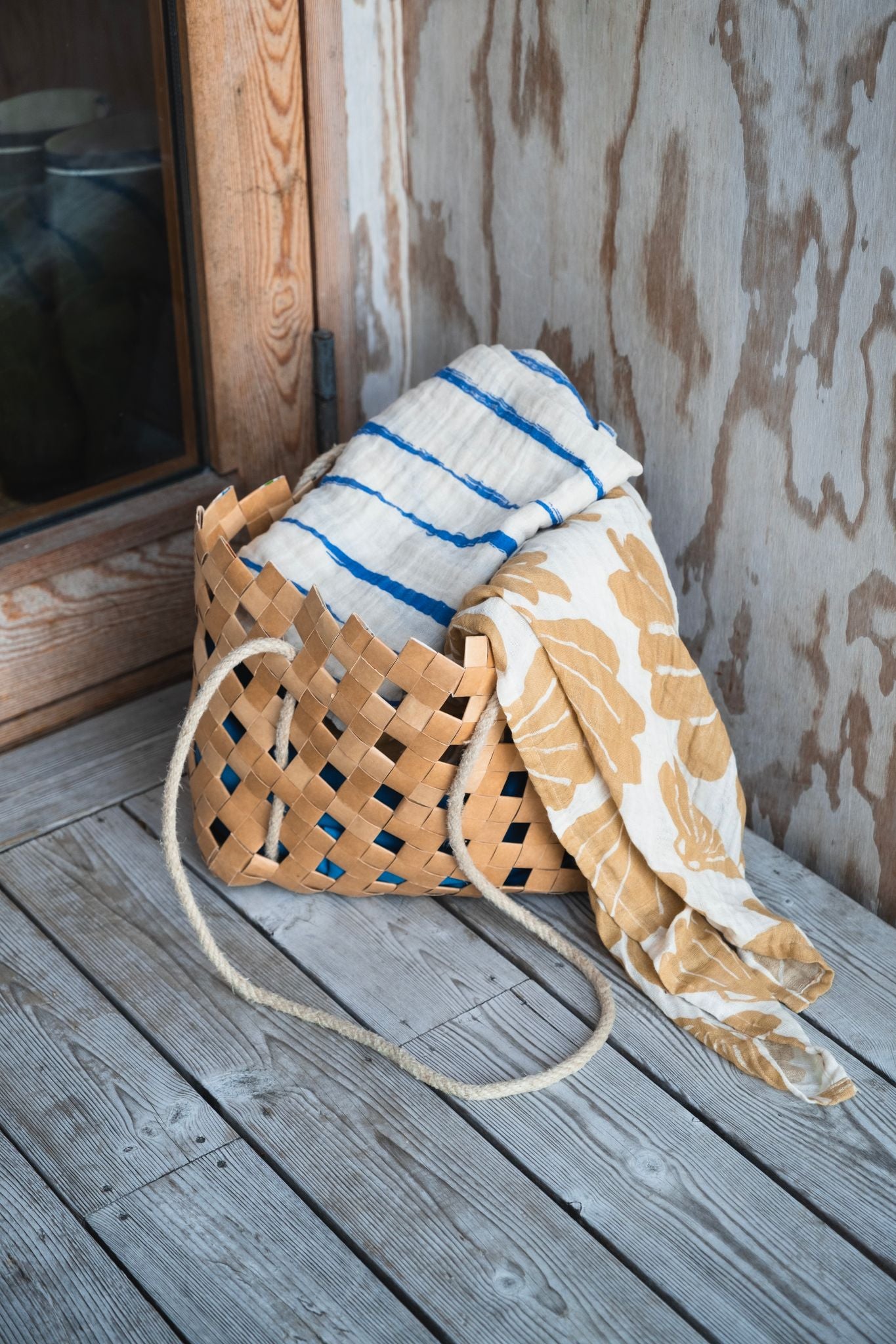 Two muslin clothes folded up in a woven bag on a wooden floor.