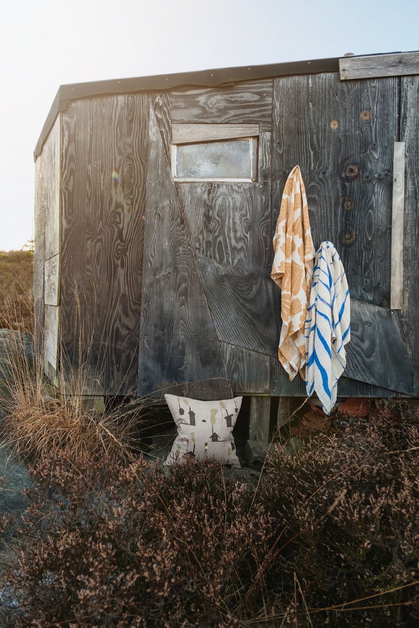 Two muslin clothes hanging off a wooden shed outside, with a cushion placed nearby on the floor.