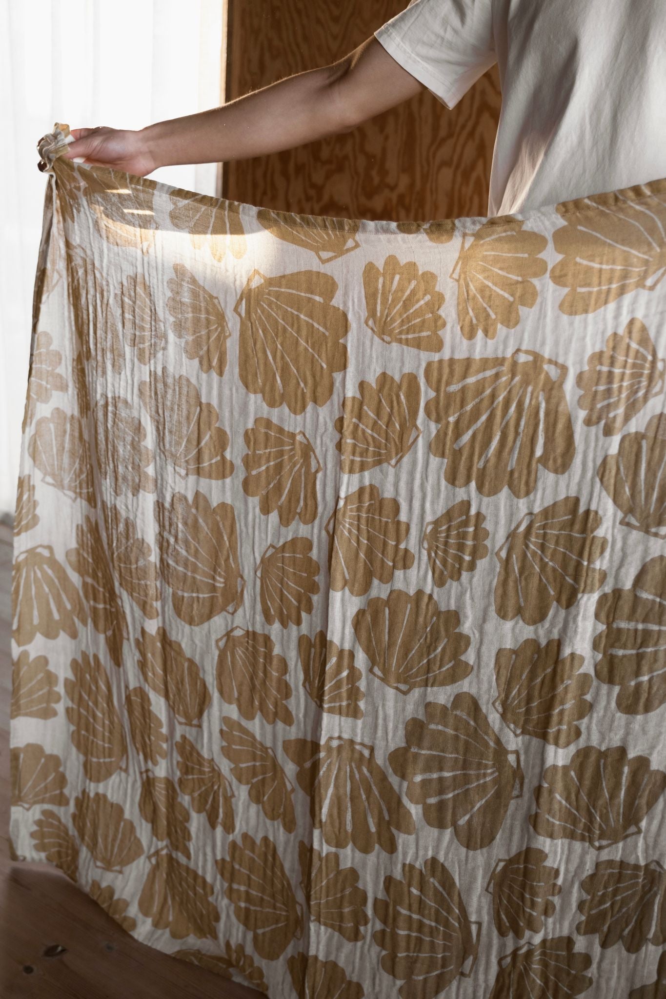 A person holding up an outstretched muslin blanket with a shell pattern in an off-white/sand colour.