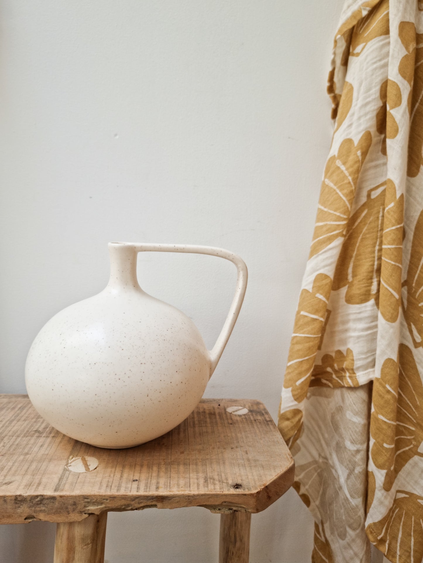 A hanging muslin blanket with a shell pattern in an off-white/sand colour next to a white vase sitting on a wooden table.