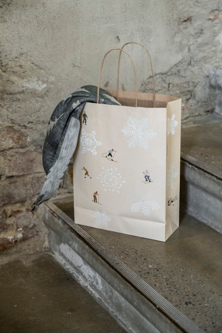 A beige paper gift bag with a skiing and snowflake pattern, featuring two handles, resting on a concrete step with a grey background.