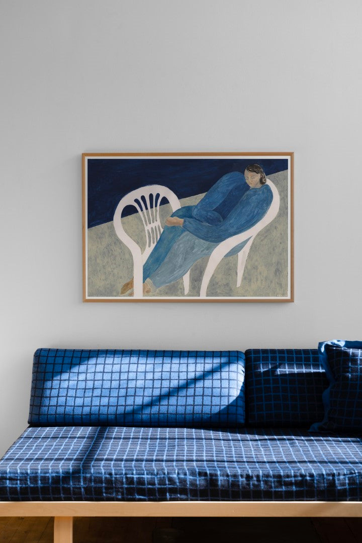 A poster featuring a blue figure lounging on a white chair, displayed on a wall above a sofa, with a frame on the wall to its left.