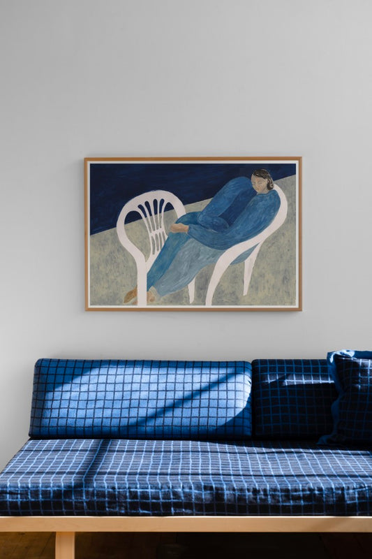 A poster featuring a blue figure lounging on a white chair, displayed on a wall above a sofa, with a frame on the wall to its left.