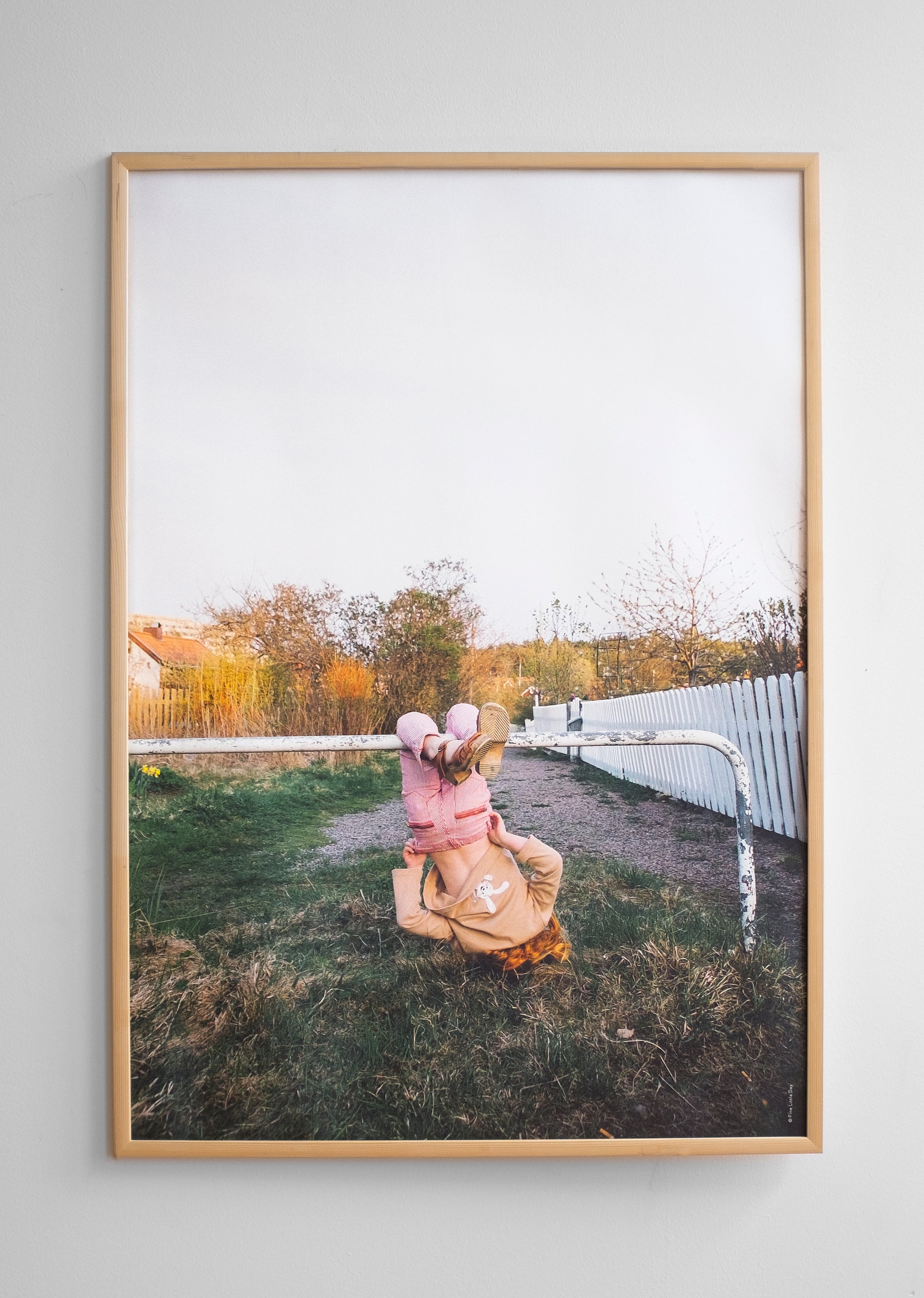 A framed poster featuring a child swinging upside down on a horizontal pole, against a grassy ground with a wooden fence and trees in the background.
