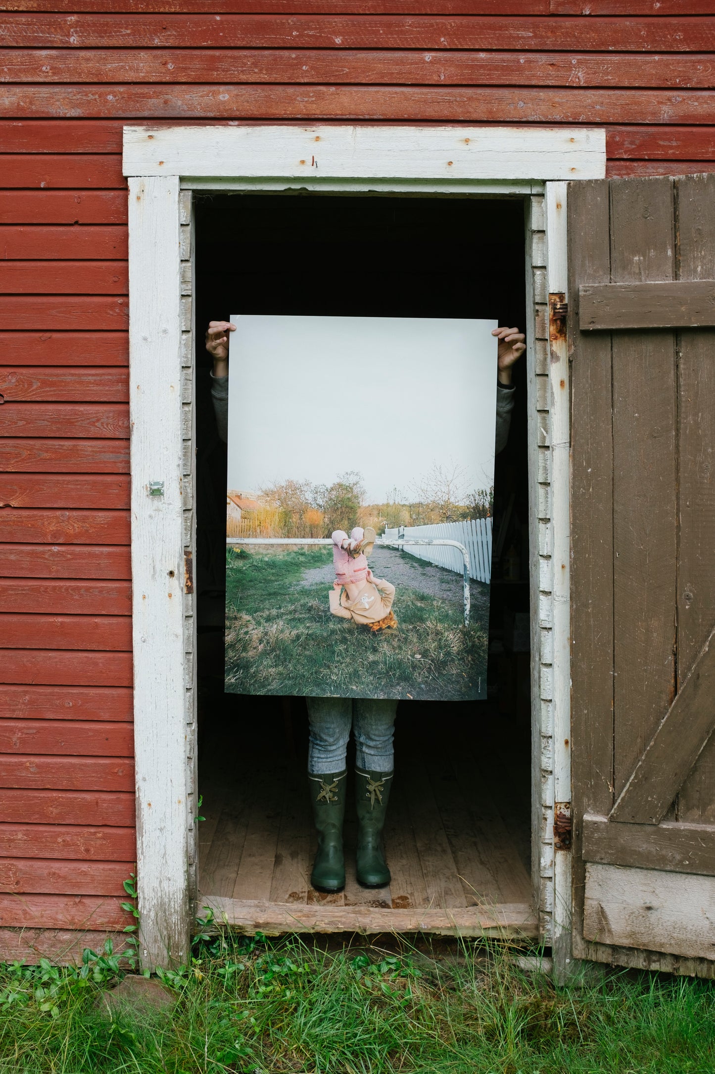 A person standing in the doorway of a building holding up a poster featuring a child swinging upside down on a horizontal pole, against a grassy ground with a wooden fence and trees in the background.