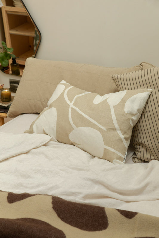 Fine Little Day Water Lilies sand cushion and Kare striped cushion against a fog linen headboard cushion on a bed with Fine Little Day Water Lilies wool blanket, organic mirror and lit candle on pine bench bedside table.
