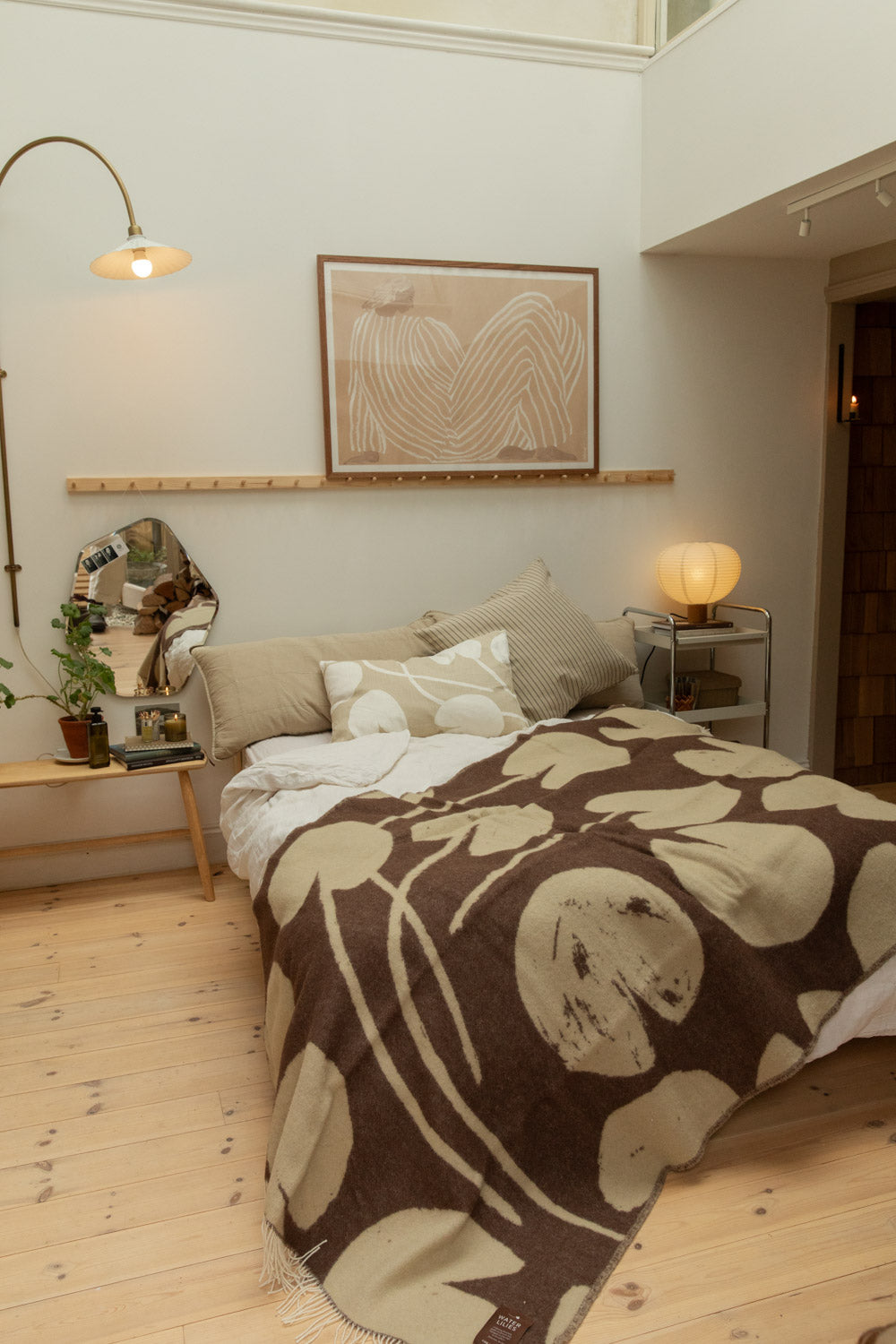 Full bedroom view with Fine Little Day Water Lilies wool blanket in chocolate brown and sand on a bed with arc brass floor lamp, abstract art print in walnut frame and pine floorboards.