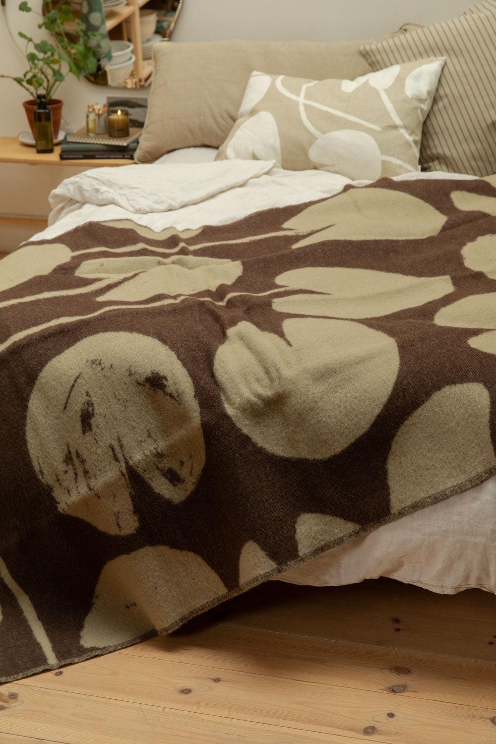 Fine Little Day Water Lilies wool blanket in chocolate brown and sand draped over a bed with lit candle, organic mirror and trailing plant on pine bench bedside table.