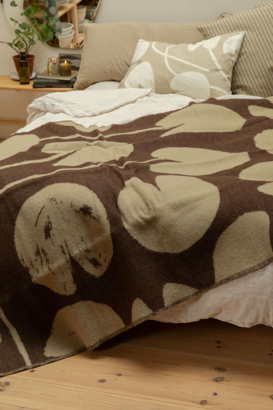 Fine Little Day Water Lilies wool blanket in chocolate brown and sand draped over a bed with lit candle, organic mirror and trailing plant on pine bench bedside table.
