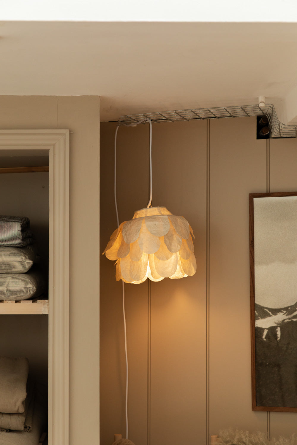 Flower petal paper pendant lampshade glowing warmly against taupe panelled wall, with folded linen on open shelving and framed print beside.