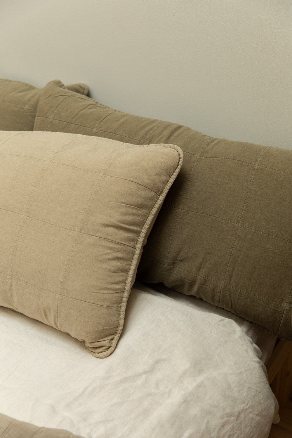 Fog and eucalyptus quilted linen headboard cushions in sand and olive green propped against each other on a white linen bed, angled view.