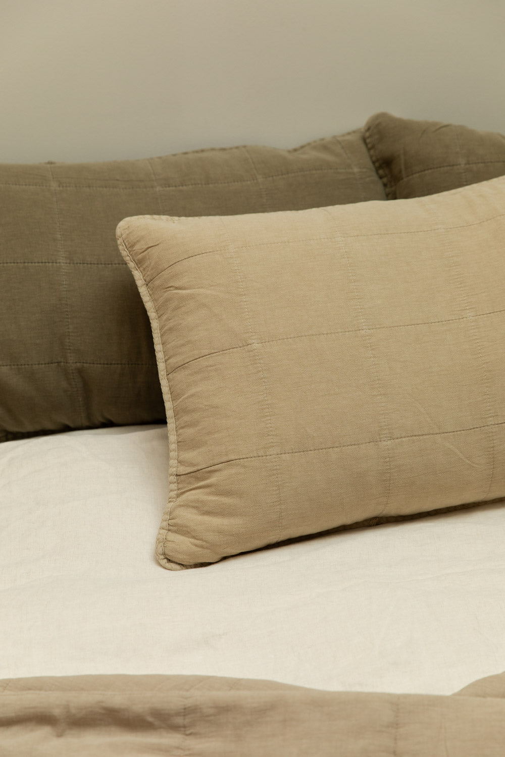 Fog quilted linen headboard cushion in sand and eucalyptus quilted linen headboard cushion in olive green styled together on a white linen bed, close-up.