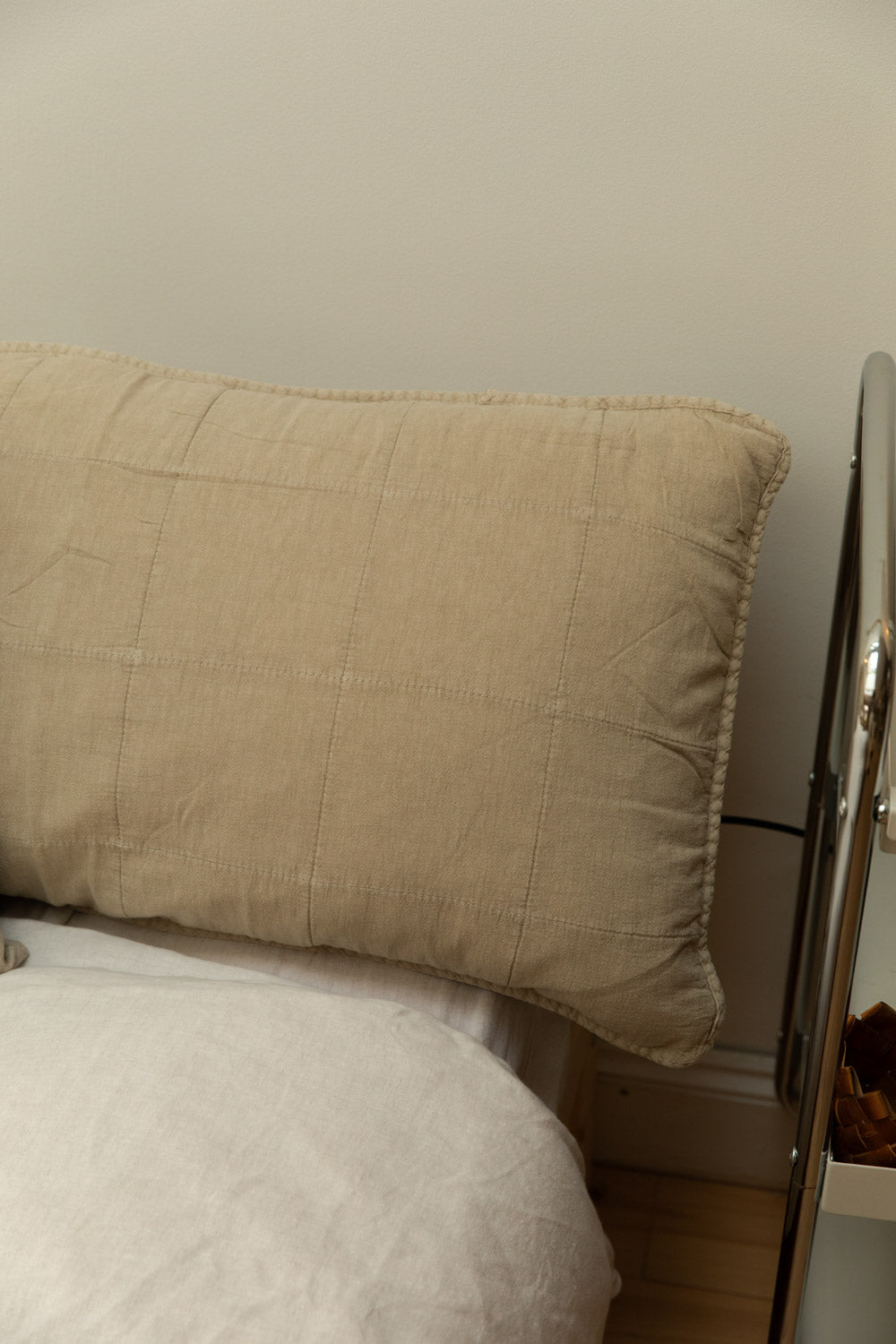 Fog quilted linen headboard cushion in sand propped against a white wall on a chrome bed frame with white linen bedding.