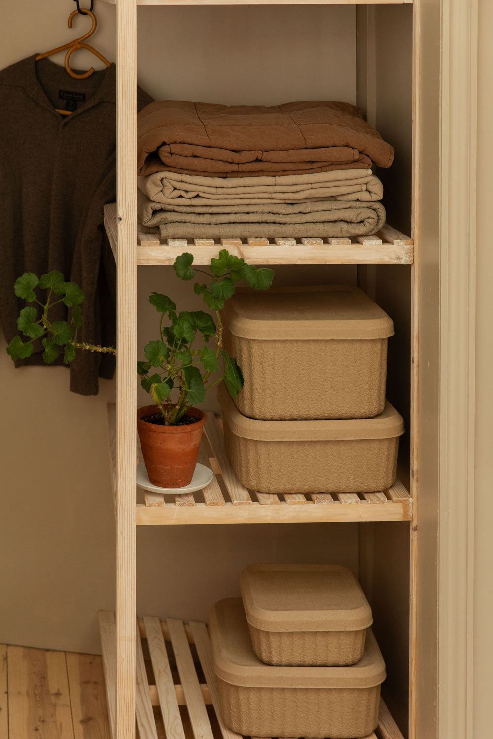 Folded terracotta and vintage linen cotton quilts stacked on open pine shelving beside Blomus Redo pulp storage boxes and a small potted geranium.