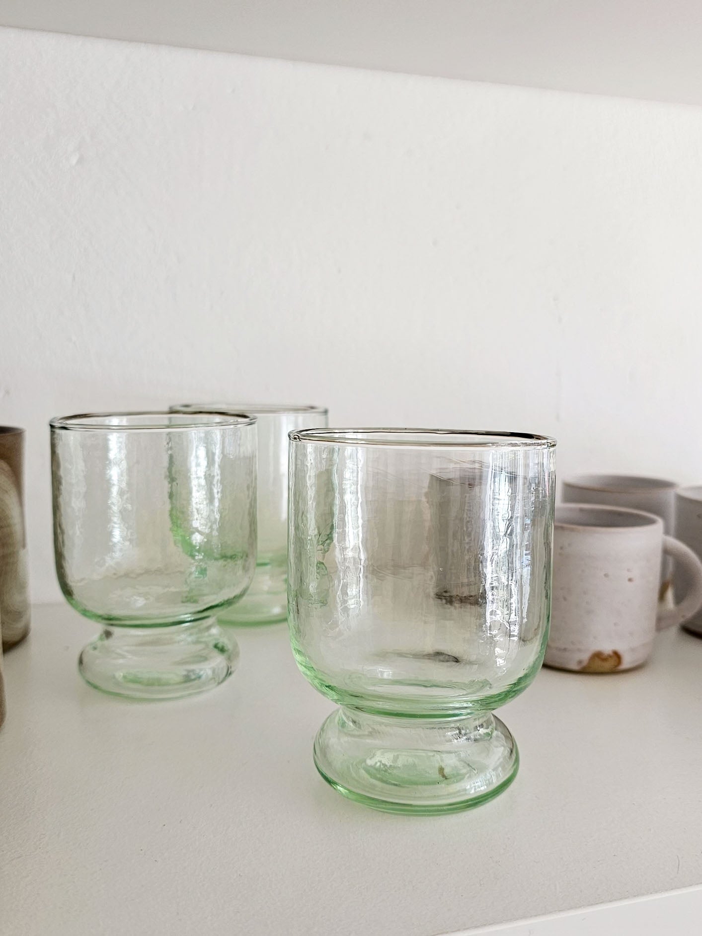 Three clear glass goblets with pale green tinted edges and pedestal bases, arranged on a white shelf against a white wall.