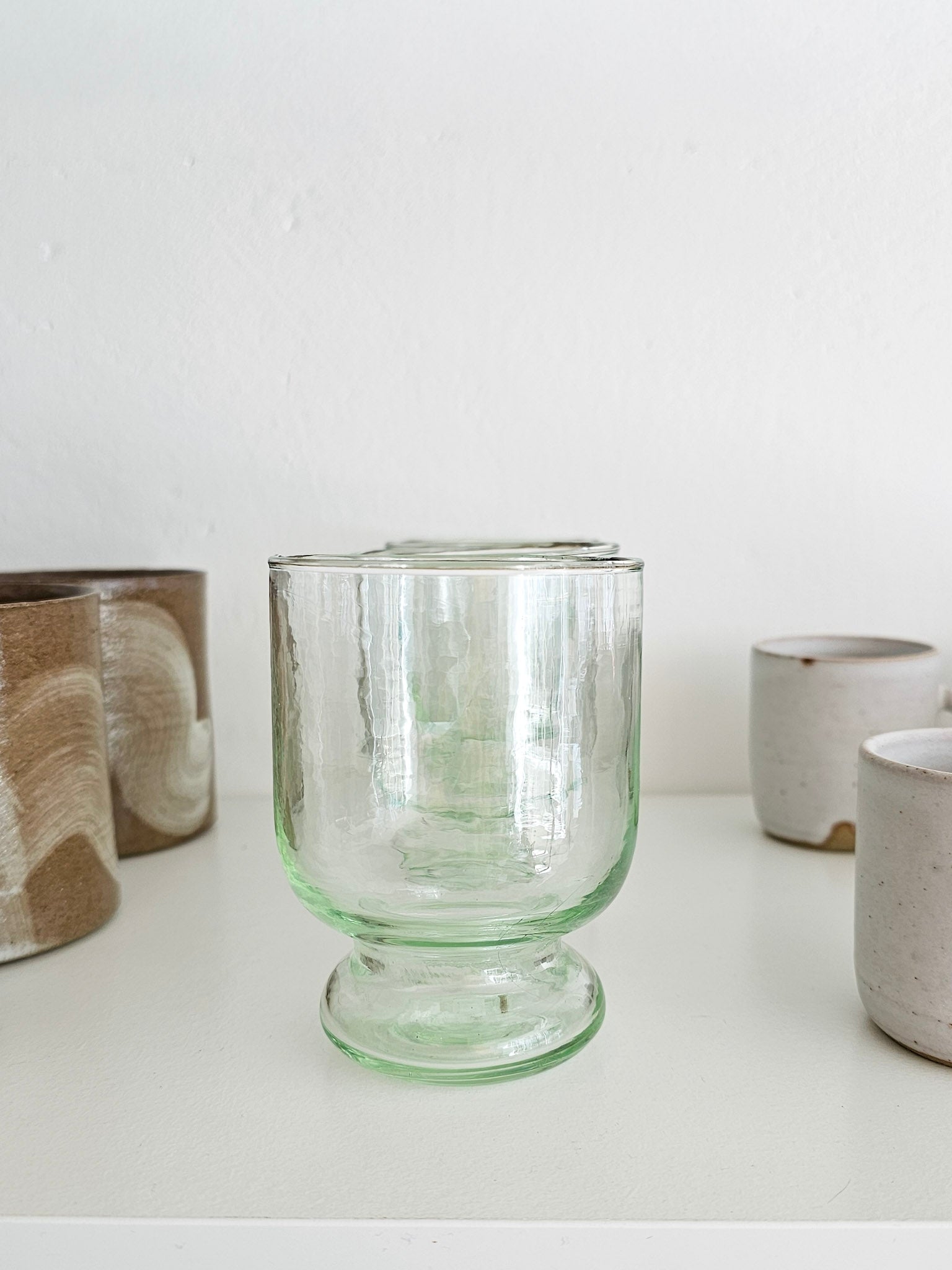 A clear light green glass tumbler with a flared base, displayed on a white surface with other ceramic cups blurred in the background.