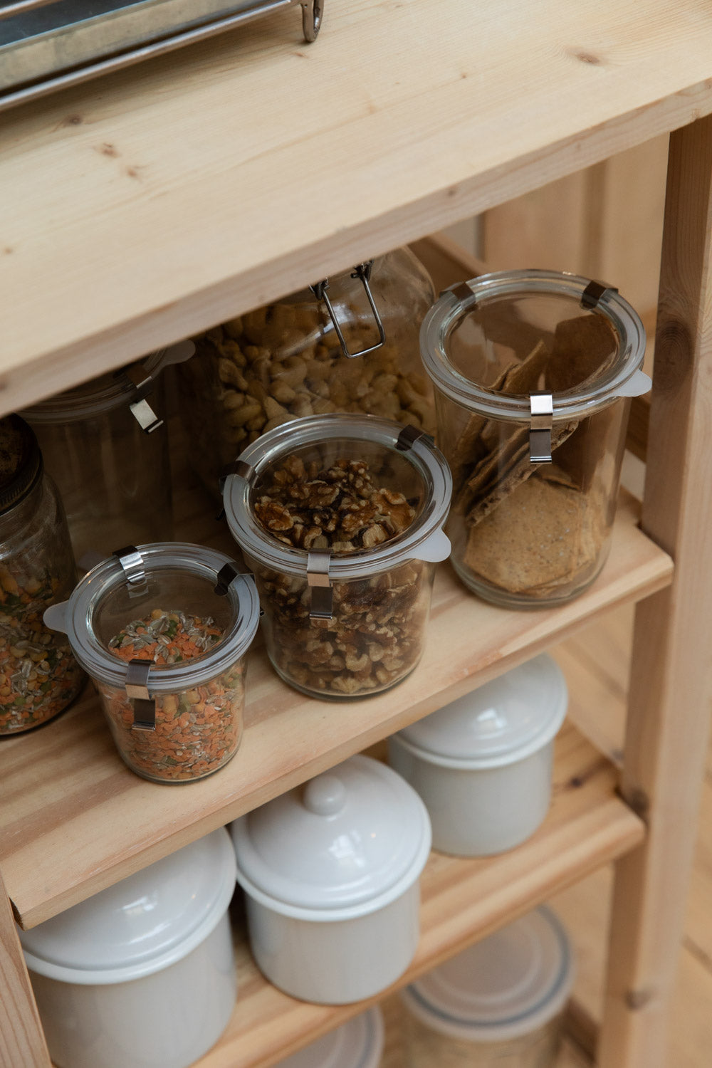 Pine shelving with glass clip-top jars of lentils, nuts and crackers above white enamel canisters