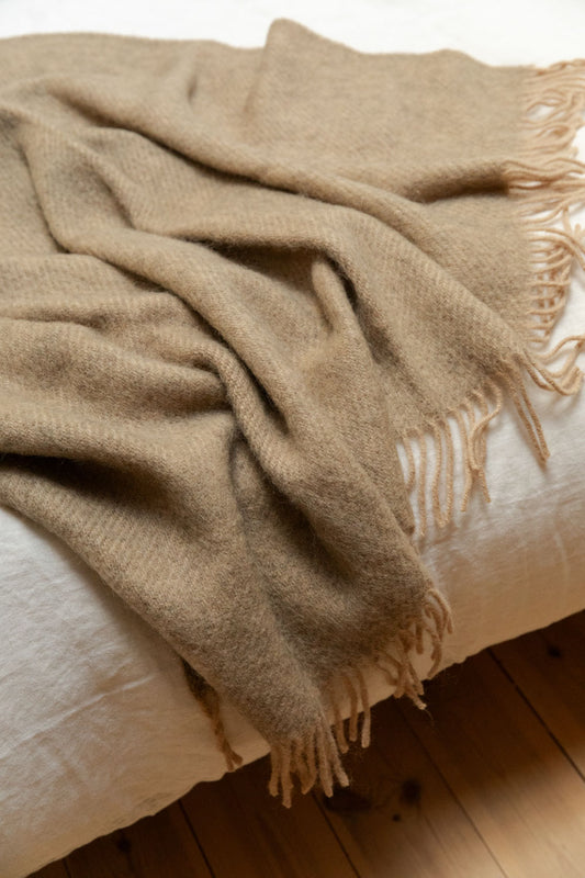 Gotland beige wool blanket with fringe detail draped over the foot of a white linen bed, showing soft herringbone weave texture.