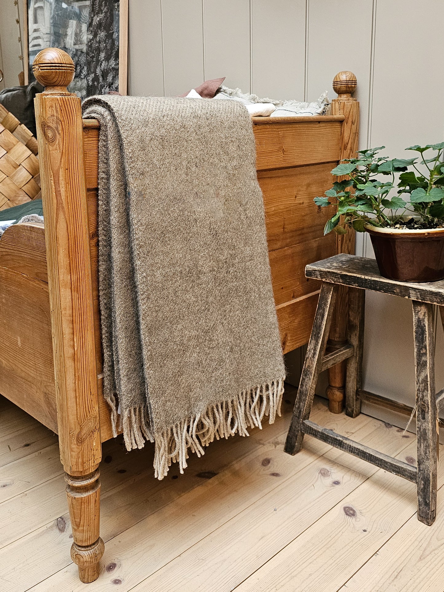 Gotland biege wool blanket draped over a wooden bed next to a wooden stool. 