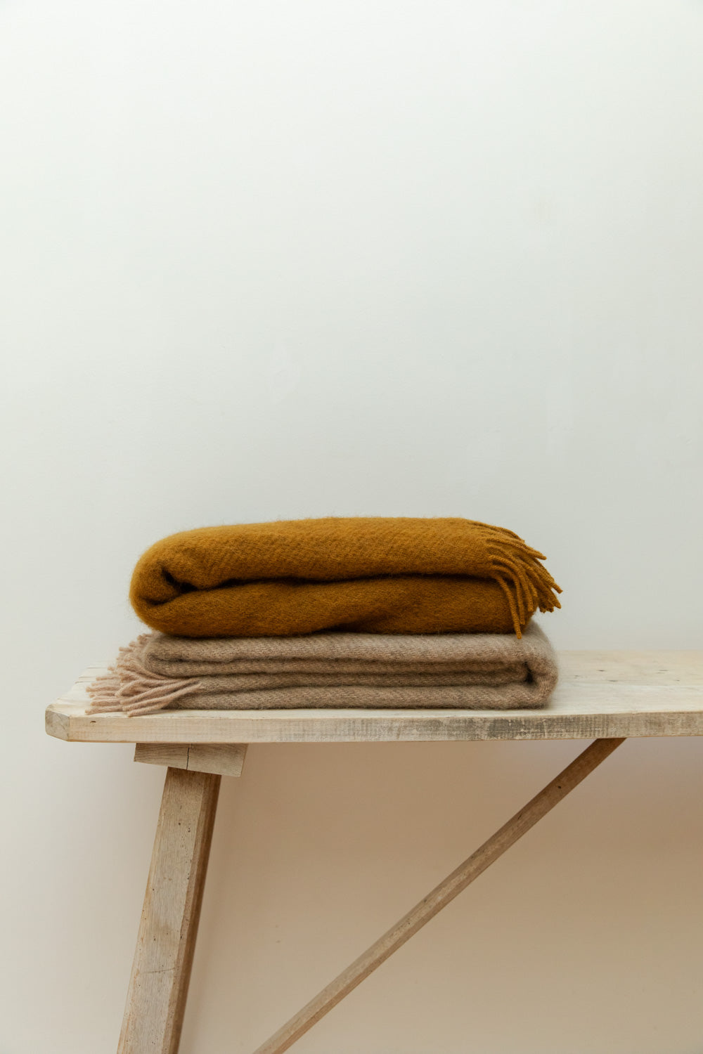 Folded Gotland caramel wool blanket with fringe detail, stacked on a pale wood trestle bench against a white wall.