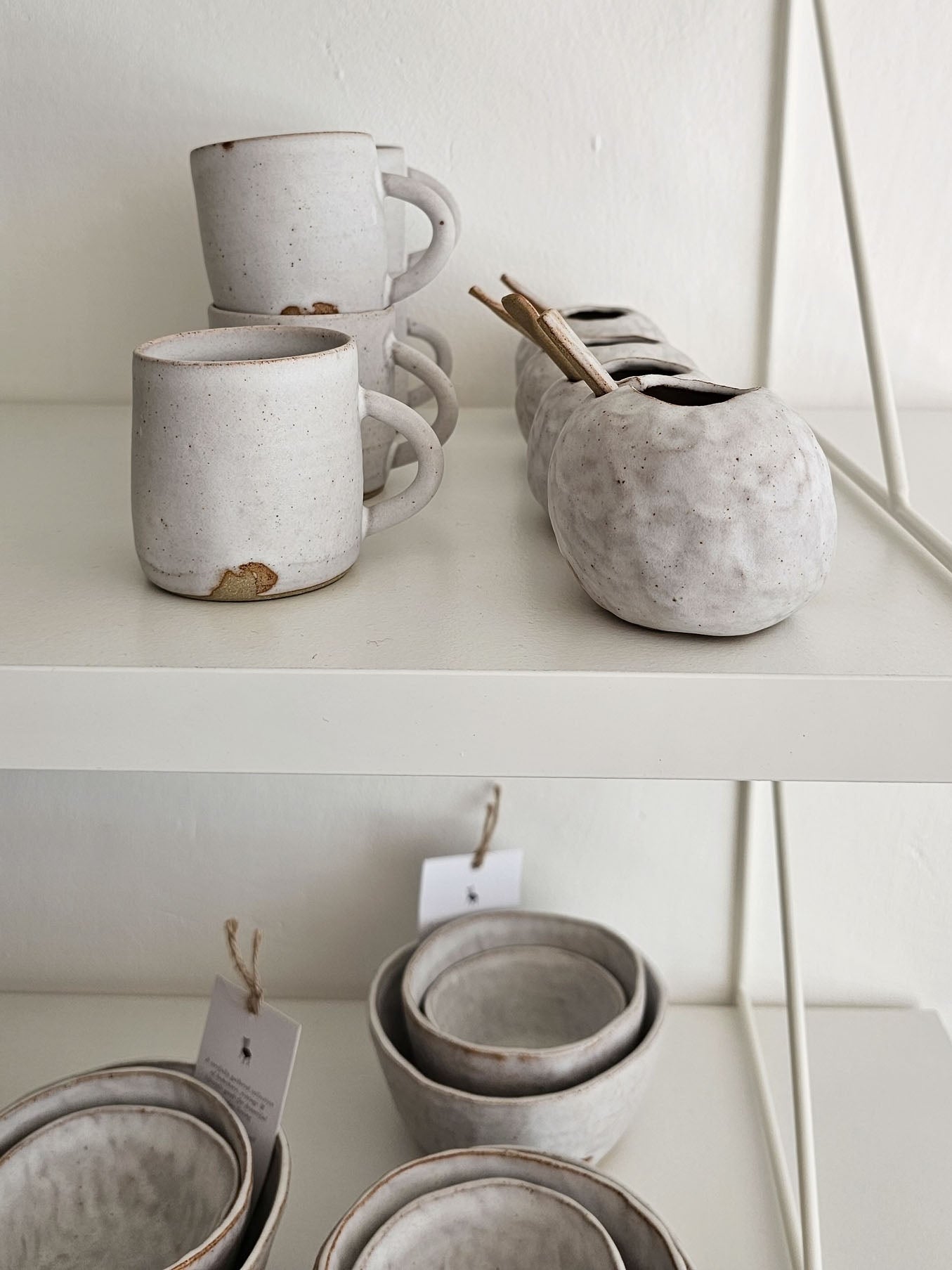 A collection of handmade, textured ceramic tableware in a light warm gray or off-white glaze, displayed on white shelves. The set includes mugs with stacked handles, a matching bowl holder with wooden utensils, and nested bowls.