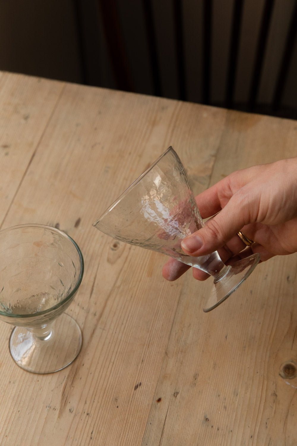 Hand lifting a hammered goblet glass beside another on a wooden table