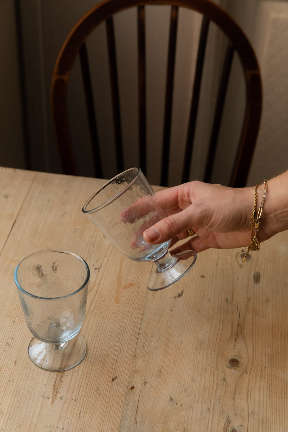 Hand picking up a hammered goblet glass from a wooden table by a spindle-back chair