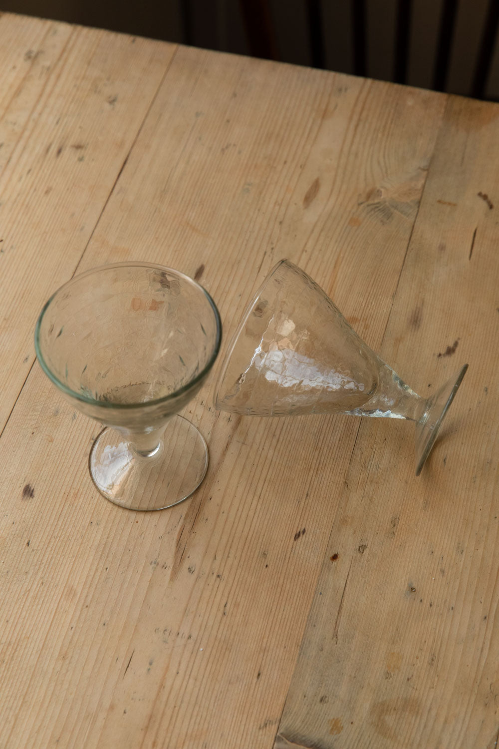 Two hammered goblet glasses on a pine table, one laid on its side
