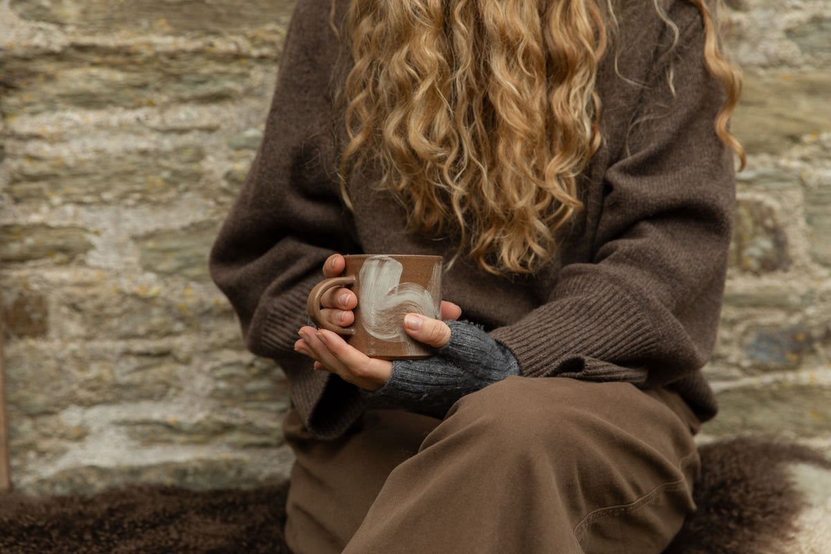 Hands in wrist warmers cradling a terracotta Brushstroke stoneware mug with white slip swirls