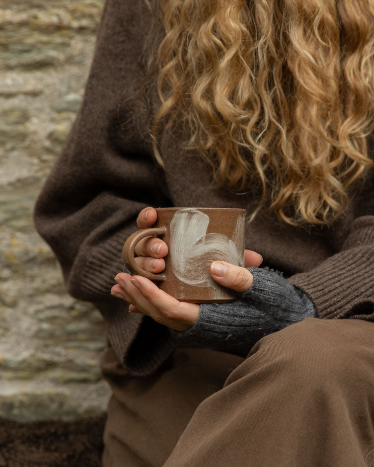 Hands in wrist warmers cradling a terracotta Brushstroke stoneware mug with white slip swirls