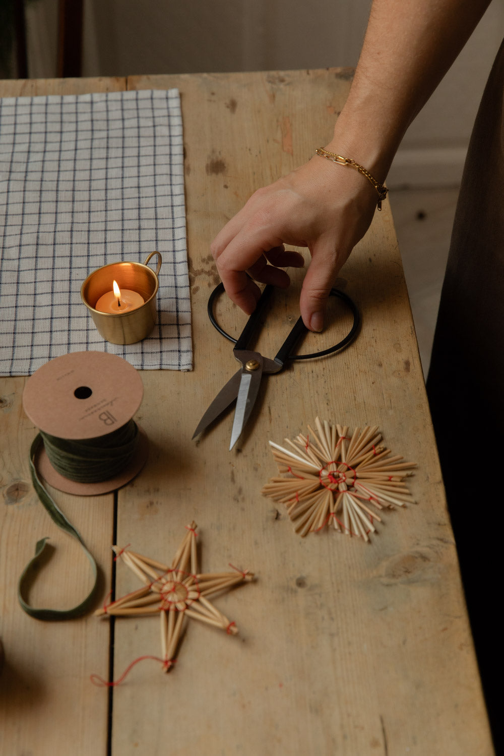 Hands with scissors among straw stars, green ribbon and a lit tealight