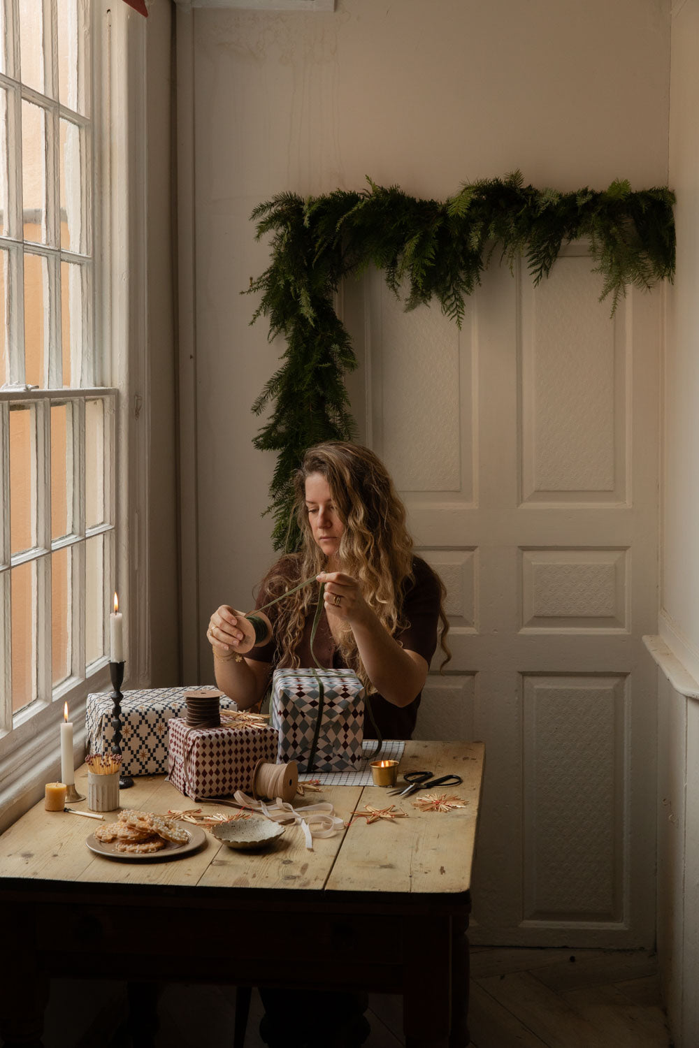 Woman wrapping presents at a kitchen table under a green foliage garland