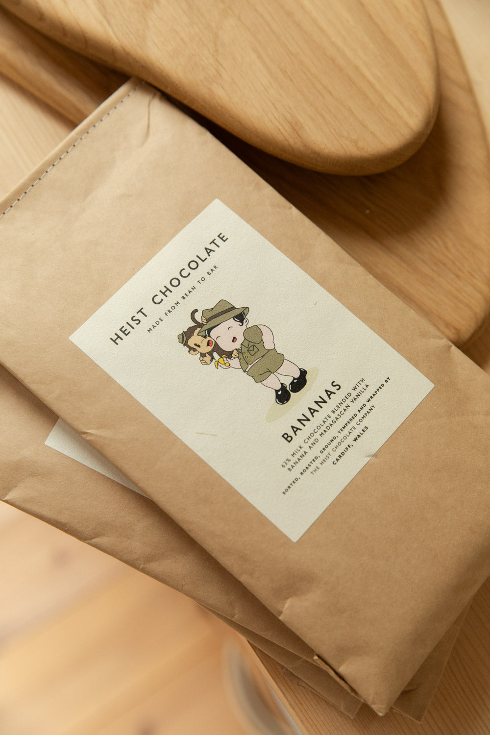 Heist Chocolate Bananas bar in kraft paper bag on a wooden surface with an oak serving board in background.