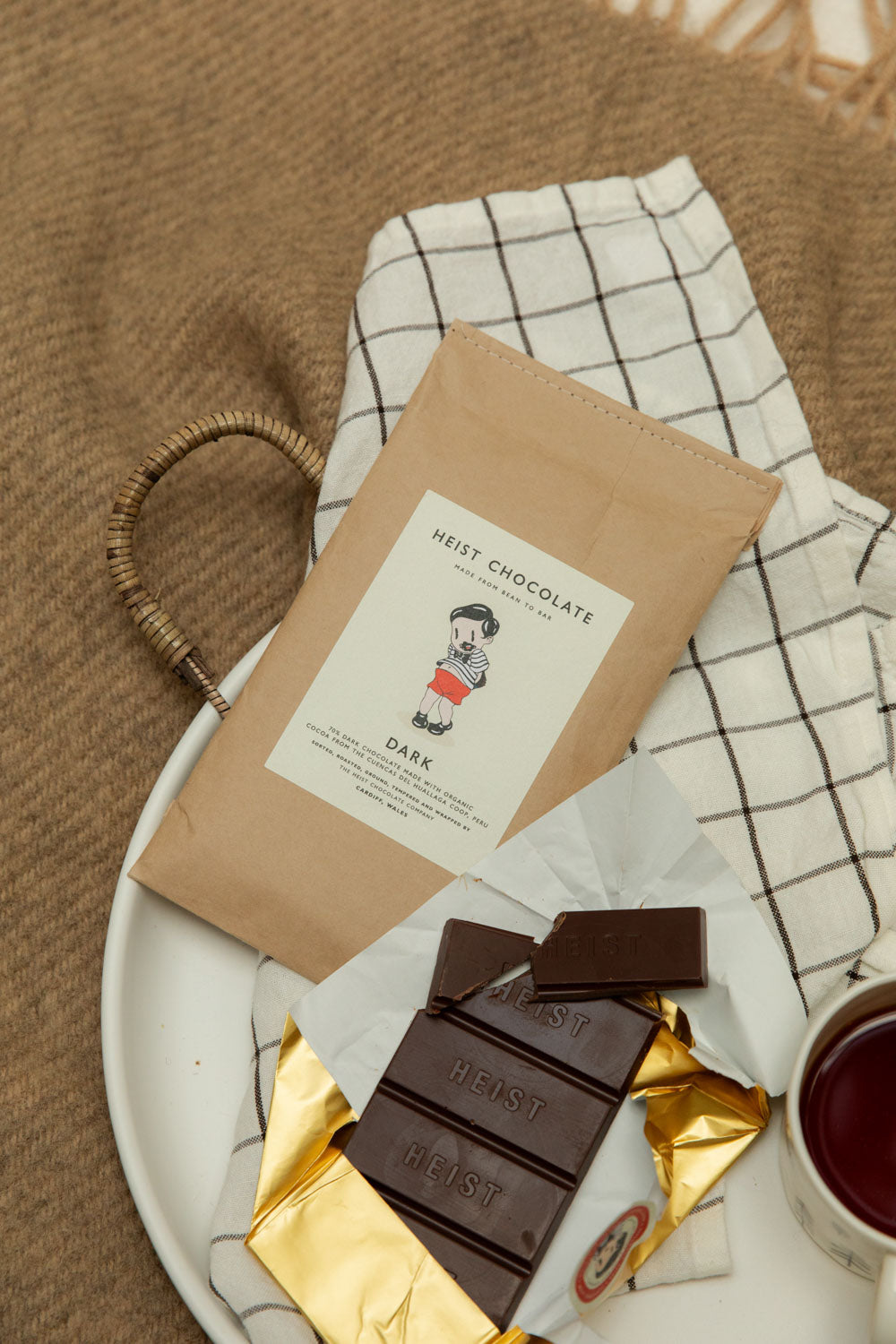 Heist Chocolate Dark 70% bar broken into pieces on a white ceramic tray with gold foil wrapping and a cup of tea.
