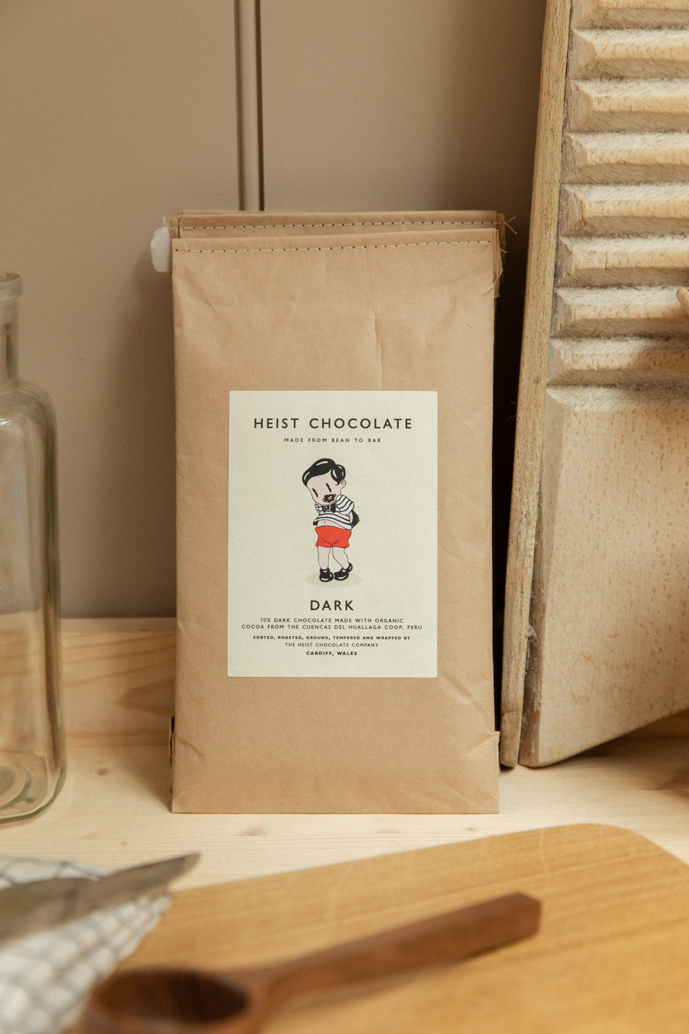 Heist Chocolate Dark 70% bar in kraft paper bag standing upright on a pale wood surface beside a washboard and glass bottle.