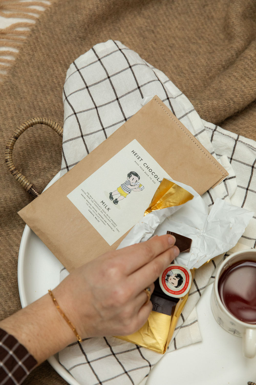Hand holding Heist Chocolate Milk bar in kraft packaging over a white tray with broken chocolate pieces and a mug of tea.