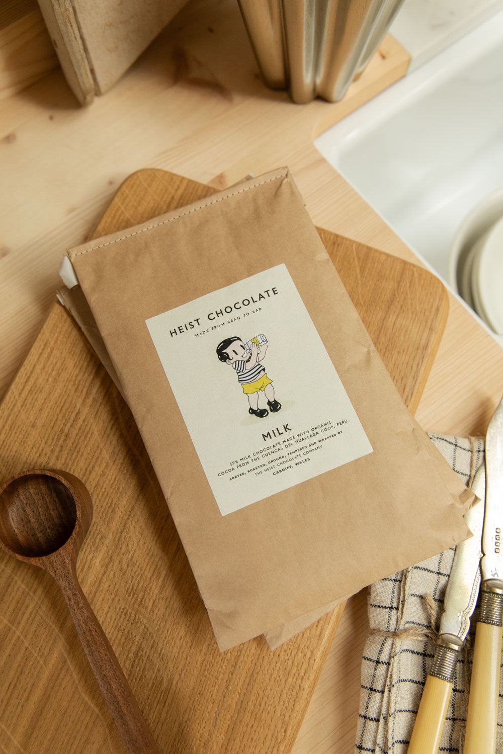 Heist Chocolate Milk bar in kraft paper packaging on an oak chopping board styled with a linen cloth and antique cutlery