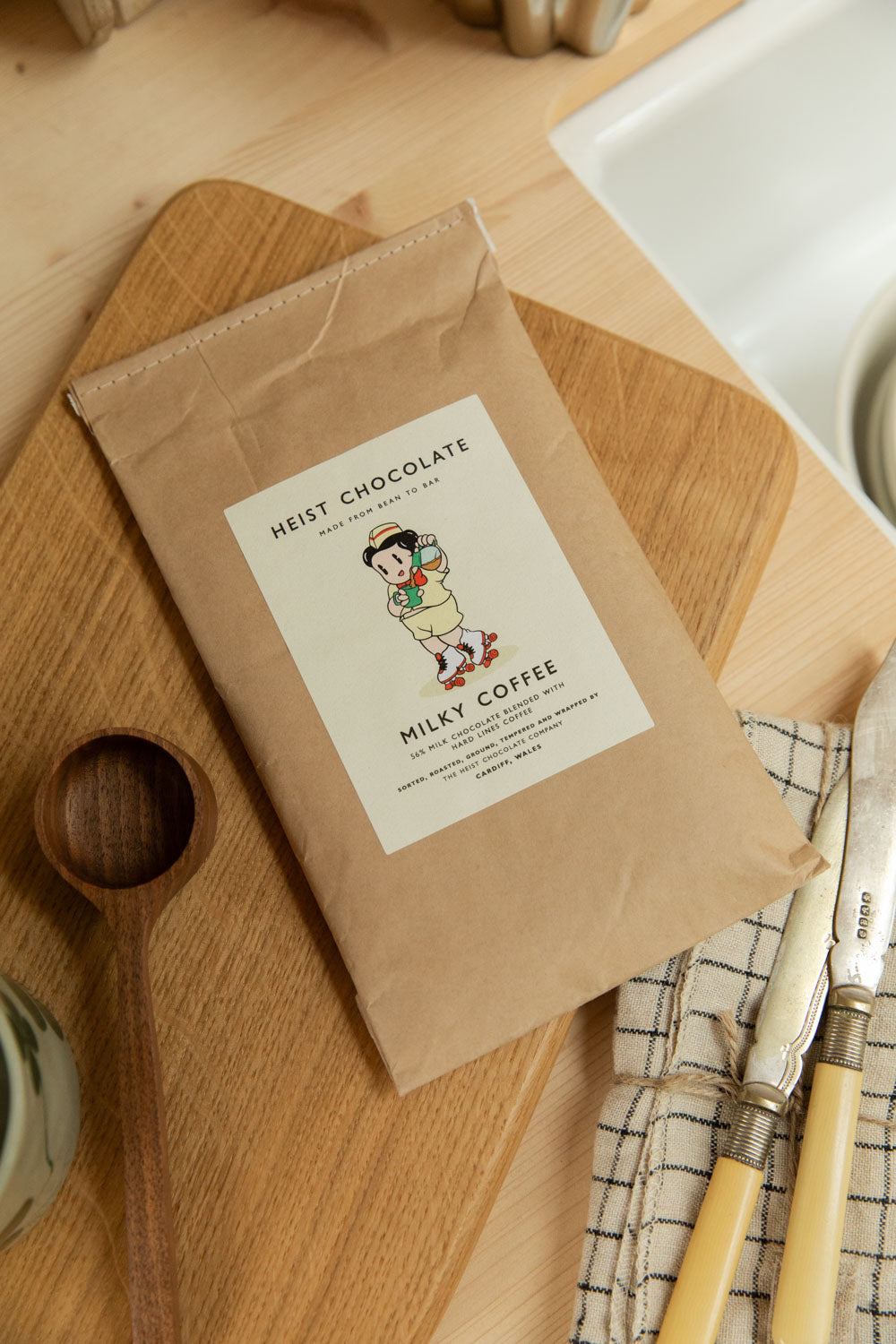 Heist Chocolate Milky Coffee bar in kraft paper packaging on an oak chopping board with a wooden spoon and linen tea towel.