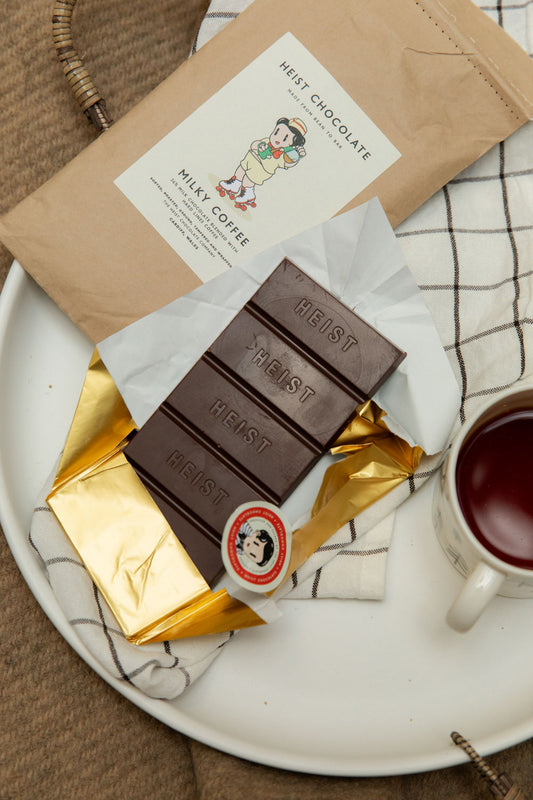 Heist Chocolate Milky Coffee bar unwrapped in gold foil on a white ceramic tray with a cup of tea.