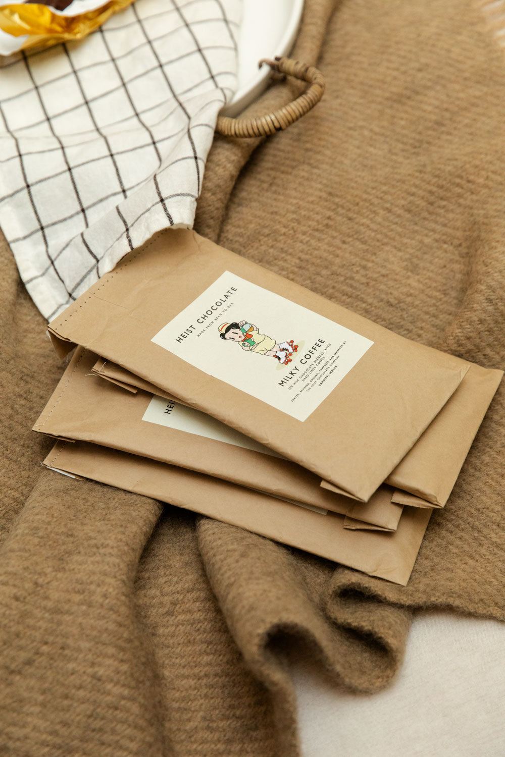 Heist Chocolate Milky Coffee bars stacked flat on a camel wool throw beside a grid linen cloth and ceramic tray.