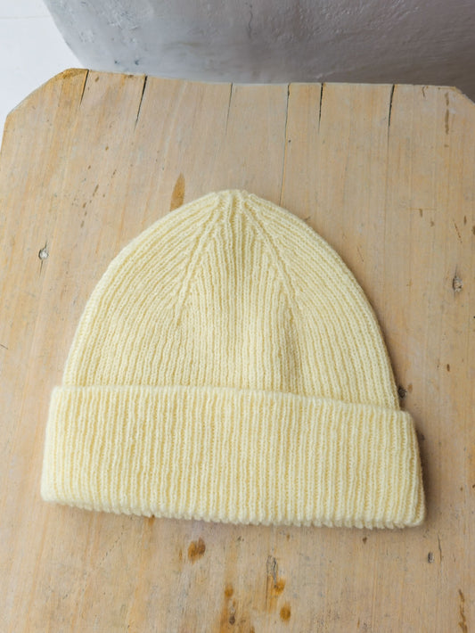 A pale ecru ribbed knit beanie hat resting on a light wood surface.