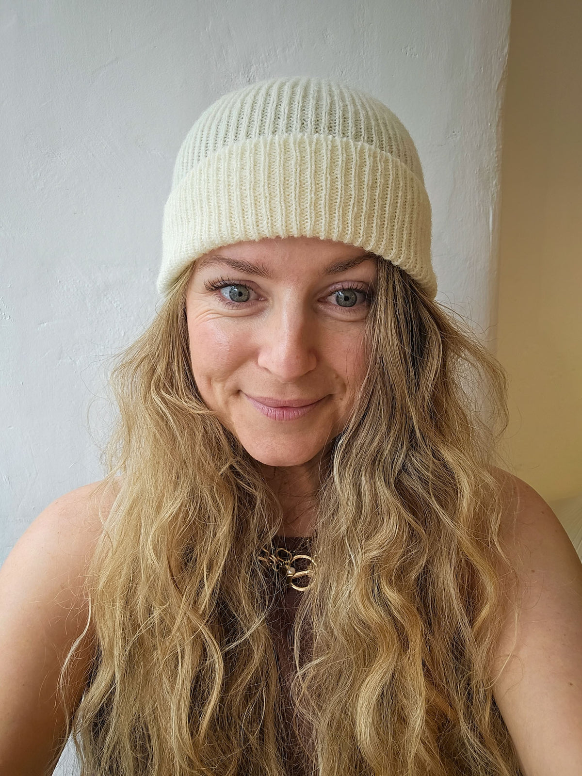 A woman with long wavy blonde hair wearing a cream-colored ribbed knit beanie hat, photographed in a close-up portrait against a neutral indoor wall.