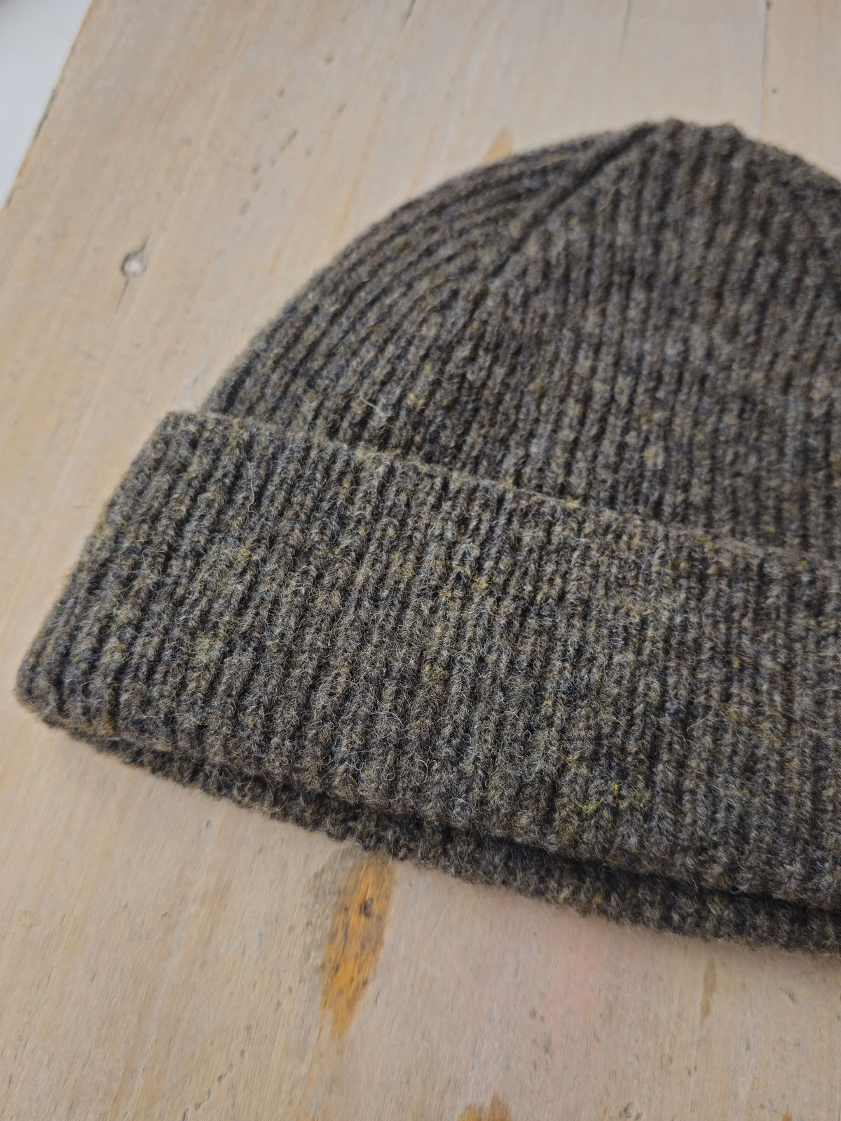 A close-up of a ribbed knit beanie in a heathered dark gray and brown tweed pattern, resting on a light wood surface.