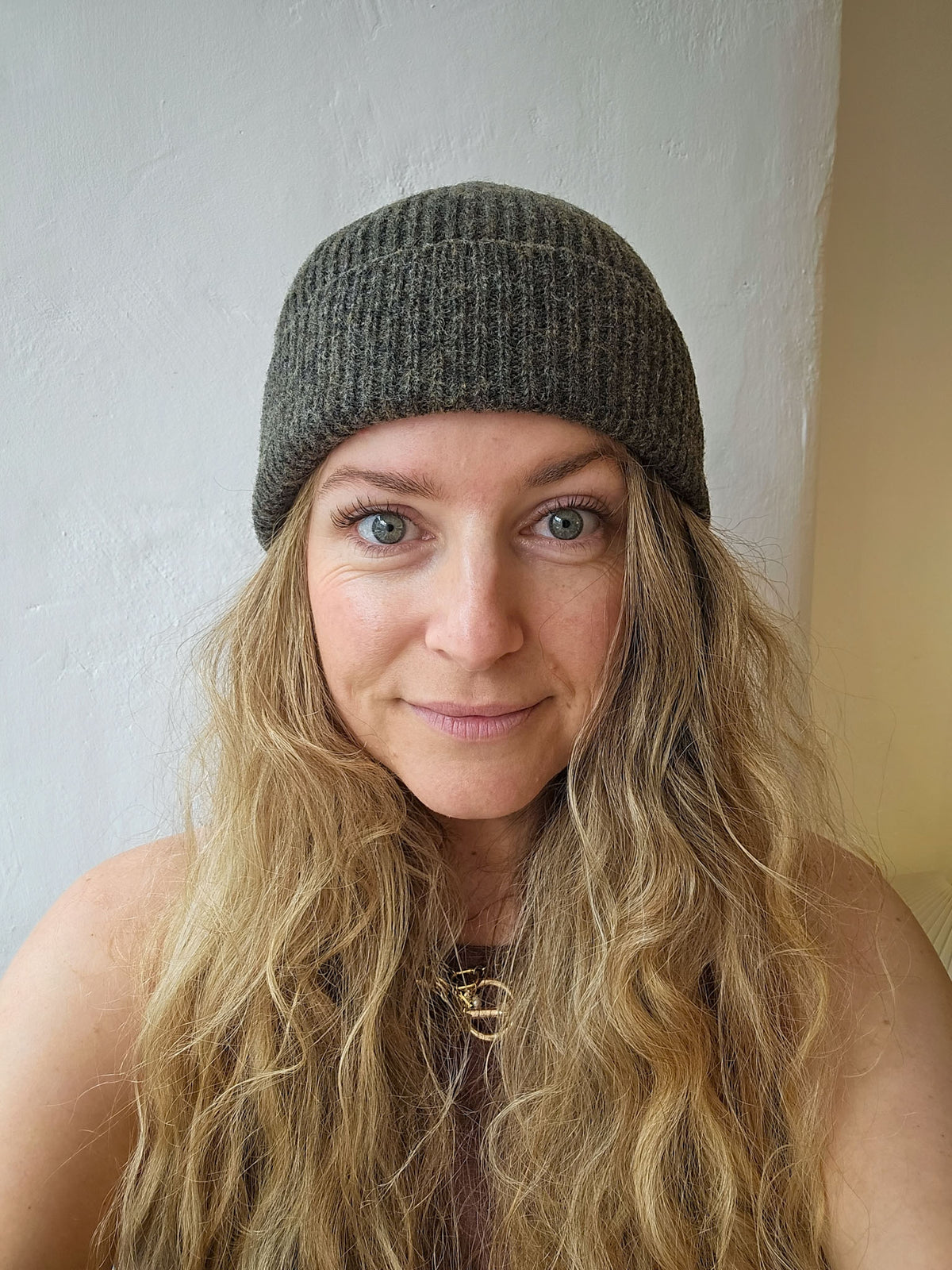 A woman with long blonde hair wearing a dark grey ribbed knit beanie hat, photographed in a close-up portrait against a light-colored wall.