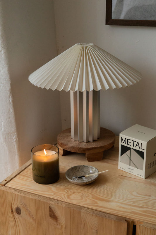 Lit candle in olive glass beside its Homework METAL box under a pleated lamp
